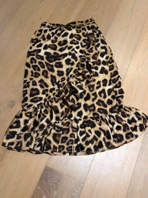 Leopardmönstrad kjol med volang - Säljer en leopardmönstrad kjol med volangkant och dekorativa rynkade detaljer framtill. Kjolen har en brun och svart färgskala och är tillverkad i ett syntetmaterial som känns lätt och smidigt. Perfekt för dig som vill sticka ut med ett djärvt mönster.