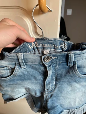 Blå jeansshorts från LAB Industries -  blå jeansshorts från LAB Industries med slitna detaljer och klassisk femficksdesign. Shortsens material är denim och de har knappstängning framtill samt bälteshällor. Perfekta för en avslappnad och trendig look.