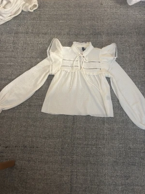 Vit blus med puffärmar och spetsdetaljer - Söt vit blus med puffiga axlar, långa ärmar och spetsdetaljer längs axlar och bröst. Blusen har knytband vid halsen och ett lätt strukturerat tyg som ger en romantisk känsla. Perfekt för dig som gillar en feminin och trendig stil.