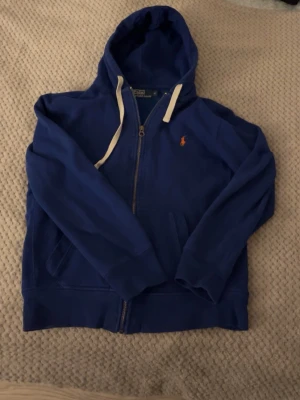 Blå zip hoodie från Polo Ralph Lauren - Snygg blå hoodie från Polo Ralph Lauren med dragkedja, loggan är orange och är broderad på bröstet. Perfekt för en chill och stilren look. Den är använd men fortfarande i väldigt bra skick. Vid frågor är det bara att skriva☺️