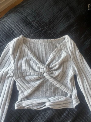 Grå ribbad topp med knyt - Säljer en grå ribbad omlottopp med långa ärmar och twistad knut framtill. Toppen har ett croppat snitt och är tillverkad i mjukt stretchigt material. Perfekt för dig som gillar trendiga detaljer och vill ha en unik topp i garderoben.