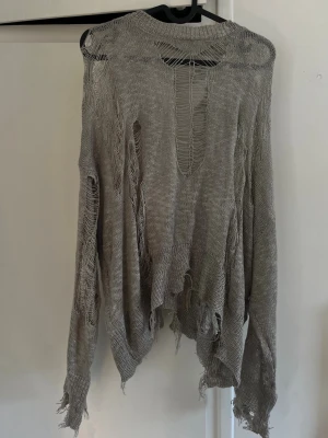 Grå distressed stickad tröja - Grå oversized stickad tröja med sliten och trasig look, stora hål och fransiga kanter. Tröjan har rund halsringning och långa ärmar. Perfekt för en edgy och alternativ stil.