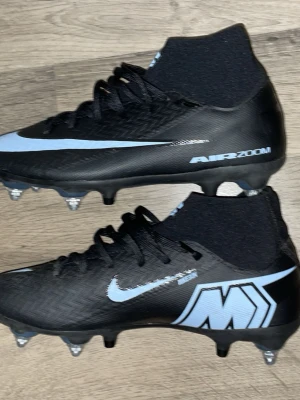 Nike Air Zoom Mercurial fotbollsskor PRO - Säljer ett par svarta Nike Air Zoom Mercurial fotbollsskor med ljusblå detaljer och logga. Skorna har strumpkrage för extra stöd runt ankeln och metallskruvdobbar för bästa grepp på planen. Tillverkade i syntetmaterial som ger bra känsla och kontroll på bollen. Zm10