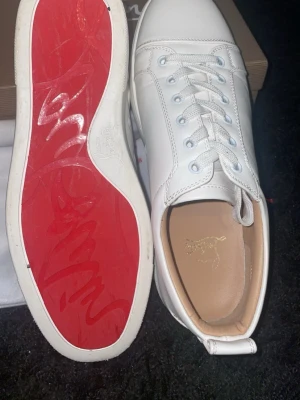 Vita sneakers från Christian Louboutin - Säljer ett par vita sneakers från Christian Louboutin med den ikoniska röda sulan och diskret logga på sidan. Skorna har klassisk låg profil, vita skosnören och är tillverkade i slätt läder med beige innersula. Perfekta för dig som gillar exklusiv och stilren design.