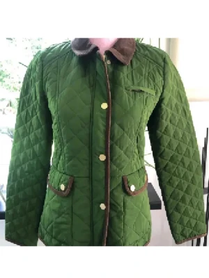 Grön quiltad vindjacka med bruna detaljer - Stilren grön quiltad vindjacka med bruna detaljer och krage i manchestertyg. Jackan har tryckknappar i guld, två framfickor med lock och dekorativa sömmar. Figurnära passform och snygga kontrastkanter ger en klassisk look.