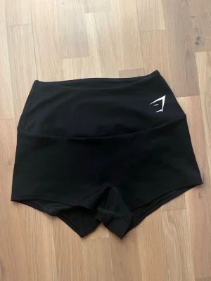 Svarta shorts från Gymshark - Säljer ett par svarta tränings shorts från Gymshark med deras logga i vitt framtill och baktill. Shortsen har en tight passform och är tillverkade i stretchigt syntetmaterial, perfekta för träning eller gym.💪