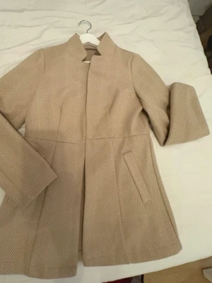 Beige kappa med fiskbensmönster - Stilren beige kappa med diskret fiskbensmönster och ståkrage. Kappan har två snedställda fickor framtill och en figurnära passform. Perfekt för dig som gillar en klassisk och tidlös look. Materialet ser ut att vara ullblandning.