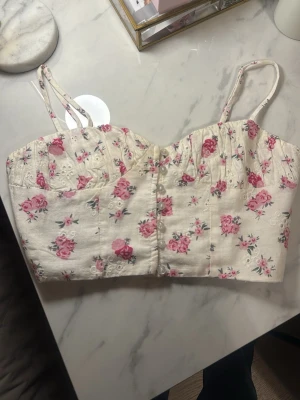 Blommig linnetopp med spetsdetaljer - Supersöt linnetopp i ljusbeige med rosa blommönster och tunna axelband. Toppen har fina spetsdetaljer upptill och små dekorativa knappar framtill. Strl S - Zara