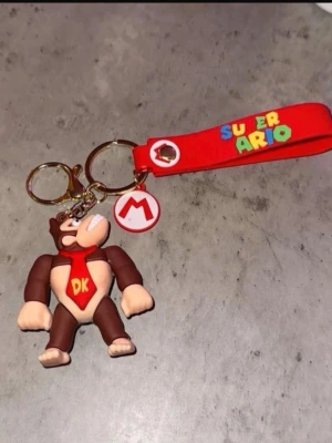 Super Mario Donkey Kong nyckelring - Nyckelring med Donkey Kong-figur i brunt och beige gummi, röd scarf med DK-logga och röd rem med färgglad Super Mario-text. Perfekt accessoar för Nintendo-fans som vill pimpa sina nycklar eller väskor.