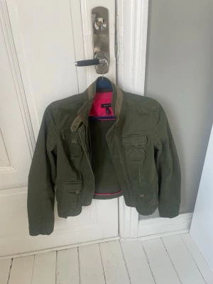 Grön utilityjacka från Gant - Snygg militärgrön utilityjacka från Gant med flera fickor framtill och coola detaljer. Jackan har en klassisk krage och dragkedja samt knappstängning. Insidan har en kontrasterande rosa detalj. Perfekt till våren/sommaren 