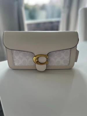 Vit axelremsväska från Coach - Snygg vit handväska från Coach med guldfärgat C-spänne framtill. Väskan har både slätt och mönstrat material med Coach-logga, samt avtagbar och justerbar axelrem. Perfekt för dig som gillar stilrena accessoarer.