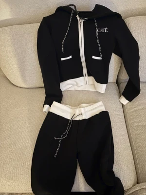 Svart och vit loungewear-set med dragkedja - Snyggt svart loungewear-set från Ceié med vita detaljer. Setet består av en hoodie med dragkedja, kedjedetaljer på snörena och två fickor samt matchande byxor med vit midja och kedjesnöre. Perfekt för chill dagar hemma.