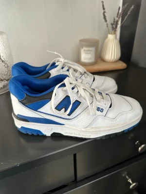 New Balance vita och blå sneakers - Säljer ett par vita och blå New Balance sneakers med svarta detaljer. Skorna har lågt skaft, klassisk siluett och är tillverkade i läder och textil. Snygg blå logga på sidan och bekväm vadderad insida. Perfekta för dig som gillar sportig stil.