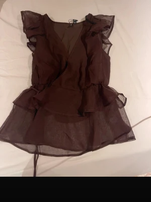 Brun omlottopp med volanger - Snygg brun omlottopp i transparent material med volangdetaljer och v-ringning. Toppen har ärmlös design och knytband i midjan för en avsmalnande siluett. Perfekt för dig som vill ha en luftig och festlig top. Storleken är satt till xs men jag skulle säga att den är mer som en S