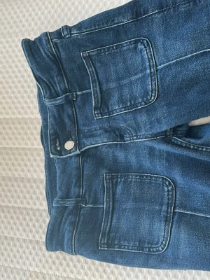 Mörkblå jeans med stora fickor - Snygga mörkblå jeans med raka ben och stora fyrkantiga fickor fram. Klassisk femficksmodell med kontrastsömmar och hög midja. Jeansen har en knappstängning och är tillverkade i ett mjukt denimtyg som ger en avslappnad look.