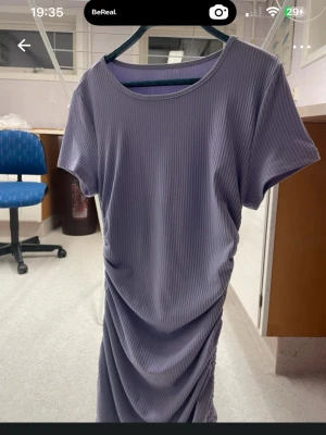 Ljuslila ribbad kortklänning - En ljuslila ribbad kortklänning med rund halsringning och korta ärmar. Klänningen har en draperad effekt på sidorna som ger en snygg och modern look. Perfekt för dig som gillar stilrena och bekväma plagg.