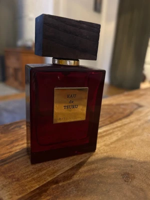 Eau de Tsuru parfym från Rituals - Elegant parfymflaska i mörkrött glas med en fyrkantig, träliknande kork och guldfärgad etikett. Doften heter Eau de Tsuru och kommer från Rituals, perfekt för dig som gillar exklusiva och stilrena parfymer. Halvfull