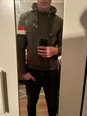 grön hoodie från A.C.E - Olivgrön hoodie med broderad logga på bröstet och färgblock på vänster ärm i rött, vitt och mörkgrönt. Justerbar huva med snören som har röda och vita detaljer. Ribbad mudd vid ärmslut och nederkant. Materialet är mjukt och bekvämt, perfekt för chill dagar. Han på bilden är 180 och väger 80kg