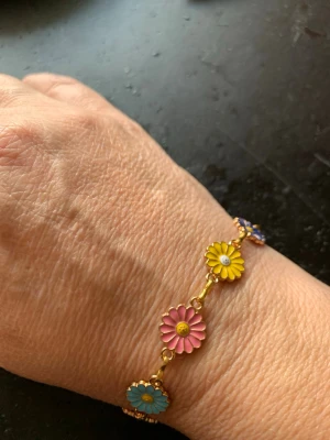 Färgglatt armband med blommor - Säljer ett armband med guldtonad kedja och emaljerade blommor i olika färger som rosa, gul, blå, röd, grön, lila och vit. Blommorna är platta och sitter tätt längs hela armbandet. Perfekt för dig som gillar färg och lekfull stil!