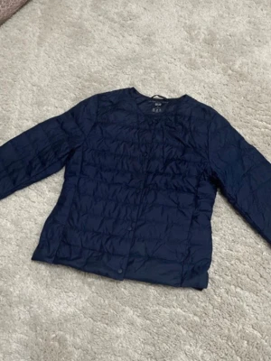 Mörkblå lättviktsdunjacka Uniqlo - Snygg mörkblå lättviktsdunjacka från Uniqlo med rund halsringning och tryckknappar framtill. Västen har en quiltad design och är tillverkad i tunt, smidigt material som passar perfekt för lager-på-lager. Enkel och stilren look.