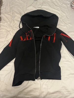 Svart zip up hoodie med röd logga från Valentino - Snygg svart zip up hoodie med dragkedja från Valentino. Stor röd logotyp tryckt över bröstet och ärmarna. Tröjan har huva och två fickor framtill. Perfekt för en trendig och avslappnad stil.