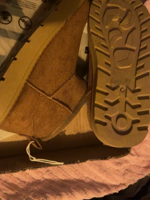 Bruna fluffiga tofflor i mocka - Mysiga bruna tofflor i mocka med vit fluffig insida. De har en robust sula med bra grepp och en enkel slip-in design. Perfekta för att hålla fötterna varma hemma. Klassisk look med mjukt material och diskret söm längs sidorna.