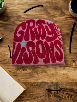 Groovy Visions rosa mössa - Säljer en rosa mössa med coolt röd texttryck 'Groovy Visions' och blå stjärnor på framsidan. Baksidan har ett rött spindelnätsmönster. Mössan är stickad i mjukt material och har en avslappnad passform. Perfekt för dig som gillar unika accessoarer med retrovibbar.