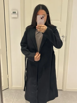 Svart lång kappa med skärp - Stilren svart kappa med klassisk krage och knytband i midjan. Kappan har långa ärmar och en rak, elegant passform som passar perfekt till höst och vinter. Materialet ser ut att vara ullblandning och kappan har en tidlös look.