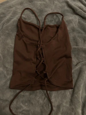Brun snörad crop top med tunna band - Trendig brun crop top med tunna axelband och snygg snörning framtill. Toppen har en åtsittande passform och är tillverkad i ett stretchigt material som känns mjukt mot huden. Perfekt för att skapa en cool och avslappnad look.
