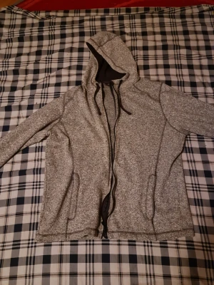 Gråmelerad hoodie med dragkedja - Snygg gråmelerad hoodie med dragkedja och huva. Mjukt material och två fickor framtill. Svart innerfoder i huvan och dragsnören för justering. Perfekt för en avslappnad stil.