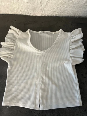 Vit ribbad topp med volangärmar - Snygg vit ribbad topp med v-ringning och korta volangärmar. Toppen har en croppad passform och är perfekt för dig som gillar en feminin och trendig stil. Materialet är mjukt och stretchigt för extra komfort.