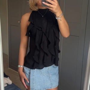 Svart volangig ärmlös blus - Supersnygg svart blus med volanger framtill och hög hals. Ärmlös modell som ger en chic och trendig vibe. Tunt, lätt material som faller snyggt och känns luftigt. Perfekt att styla med jeans eller kjol för en cool look.