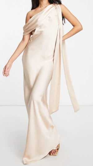 Champagnefärgad balklänning i satin - Elegant långklänning i champagnefärgad satin med oneshoulder-design och draperad detalj över axeln. Klänningen har en mjuk, glansig yta och en figurnära passform som ger en lyxig känsla. Perfekt för dig som vill sticka ut med en stilren look. Använt endast 1 gång 