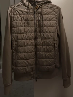 Beige pufferjacka från Parajumpers - Snygg beige pufferjacka från Parajumpers med quiltad front, huva och dragkedja. Jackan har både fleece- och pufferdetaljer, praktiska fickor med dragkedja och en bröstficka. Perfekt för kalla dagar och har en modern look. Den har en liten skada på nere vid handleden! Men är inget märkvärdigt och cardiganen är äkta! 
