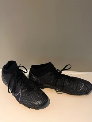 Svarta Nike Mercurial fotbollsskor - Säljer ett par svarta Nike Mercurial fotbollsskor med högt skaft och snörning. Skorna har en snygg textur i syntetmaterial och diskreta silverdetaljer, perfekt för dig som vill ha stilrena och snabba skor på planen.