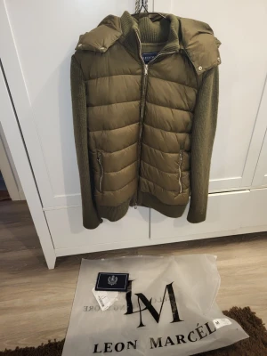 Helt ny Olivgrön cardigan från Leon Marcel  - Snygg olivgrön cardigan från Leon Marcel med stickade ärmar och avtagbar huva. har två dragkedjeförsedda fickor och ribbade muddar. Perfekt för lager på lager och ger en cool streetwear-look.