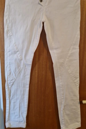 Vita jeans med detaljer från CC by Capri Collection - Säljer ett par vita jeans från CC by Capri Collection med snygga detaljer och metallknappar över vanlig dragkedje-gylf. Jeansen har smal passform och är tillverkade i vit denim med 98% bomull och 2% elastan som ger lätt stretch. Använda enstaka gång. Inga fläckar eller solkiga kanter.