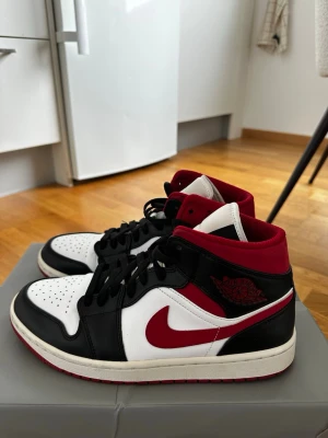 Nike Air Jordan 1 Mid röd/svart/vit - Snygga Nike Air Jordan 1 Mid sneakers i svart, vitt och rött. Klassisk design med röd swoosh, svart läderpanel och vit tåbox. Röd vadderad krage och yttersula, svart snörning och ikonisk Air Jordan-logga på sidan. Perfekta för dig som gillar streetwear och basketstil. I grmy skick.