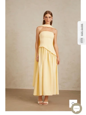 Ljusgul axelbandslös långklänning -  Ljusgul långklänning med asymmetrisk midja och axelbandslös design. Klänningen har en elegant halsdetalj och ett mjukt, flytande fall. Perfekt för dig som vill sticka ut med en modern och stilren look. Klänningen är helt ny och säljs då den kom för sent. Nypris är 2143 kr.
