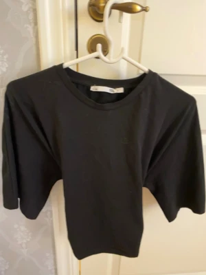 Svart topp med knyt i ryggen - Svart kortärmad topp med rund halsringning och snygg knytning i ryggen. Tillverkad i mjuk bomull, perfekt för en stilren och enkel look. Passformen är normal och toppen är enkel att matcha med olika outfits.