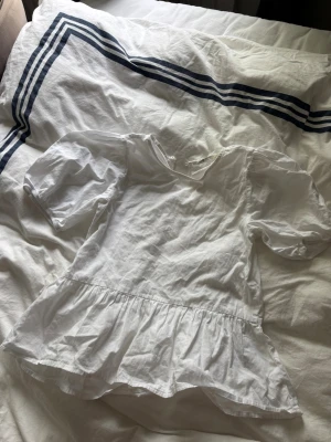 Vit blus med volang från H&M - Söt vit blus från H&M med volang nertill och korta puffärmar. Blusen har en v-ringning och är tillverkad i bomull, vilket gör den luftig och skön. Perfekt för dig som gillar en romantisk och stilren look.