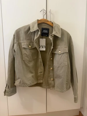 Grön jeansjacka från ZARA - Stilren grön jeansjacka från ZARA med klassisk krage, två bröstfickor med knappar och guldfärgade detaljer. Rak passform och långa ärmar med knapp vid ärmslut. Perfekt för en avslappnad och trendig look.  (Saknar en enda knapp de va så jag köpte den vid armen de kan man se på bilden) 