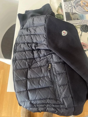 Moncler cardigan - Snygg mörkblå pufferjacka från Moncler med quiltad front och rygg i glansigt material samt stickade ärmar och krage. Två dragkedjeförsedda fickor framtill och diskret Moncler-logga på ärmen. Perfekt för kalla vinterdagar.