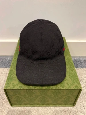 Svart keps med GG-mönster - Säljer en svart keps med det klassiska GG-monogrammet i tyget. Kepsen har justerbart spänne bak och en grön-röd rand på sidan som ger en ikonisk touch. Materialet är tyg och kepsen har böjd skärm.