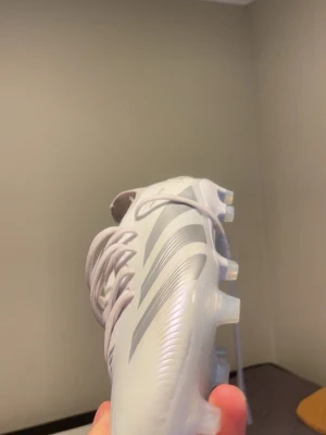 Vita Adidas fotbollsskor med silverdetaljer - Snygga vita fotbollsskor från Adidas med klassiska silverfärgade ränder på sidan. Skorna har snörning och en modern, strömlinjeformad design med fasta dobbar för spel på gräs. Tillverkade i syntetmaterial som ger bra passform och känsla på planen. Skorna heter adidas predator elit