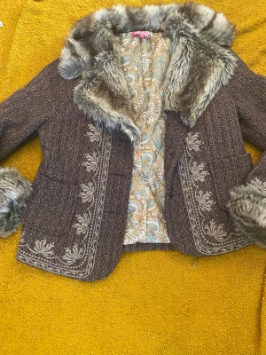 Brun kappa med fuskpäls och brodyr - Superfin brun kappa med fuskpäls vid krage och ärmslut. Snygga broderade detaljer framtill och två fickor. Insidan har paisleymönstrat foder i beige och blått. Perfekt för dig som gillar vintagevibbar och unika detaljer.