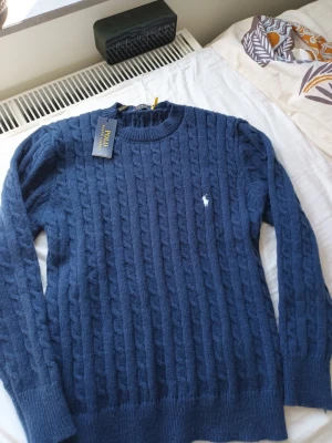 Mörkblå kabelstickad tröja Polo Ralph Lauren - Kabelstickad mörkblå tröja från Polo Ralph Lauren med rund halsringning och den klassiska vita logobroderingen på bröstet. Tröjan har långa ärmar och ribbade muddar vid ärmslut och nederkant. Perfekt för dig som gillar stilren och tidlös design.