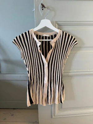 Randig ribbad topp från Zara - Säljer en ribbad topp från Zara med vertikala svarta och beige ränder. Toppen har korta ärmar, knappar framtill och är tillverkad i ett mjukt, stretchigt material. Perfekt för dig som gillar en avslappnad men ändå trendig look.