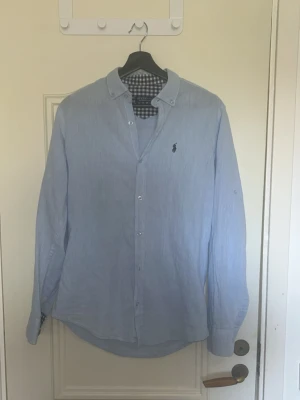 Ljusblå skjorta från Ralph Lauren - Klassisk ljusblå skjorta från Ralph Lauren med button-down krage och mörkblå broderad logga på bröstet. Skjortan har långa ärmar, knappar framtill och rutigt innerfoder i kragen. Perfekt till jeans eller chinos.