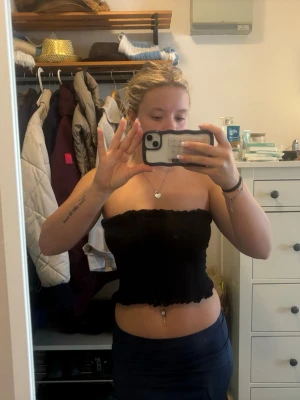 Svart bandeau tubtopp - Säljer en svart bandeau tubtopp med smockad resår och volangkant upptill och nedtill. Toppen är axelbandslös och croppad, perfekt för varma dagar eller som layering piece. Enkel att matcha med både jeans och kjol.